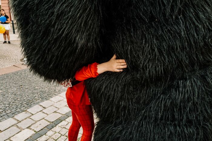 A small child in red embracing a large, shaggy black costume, a touching candid street photo for URBAN Photo Awards.