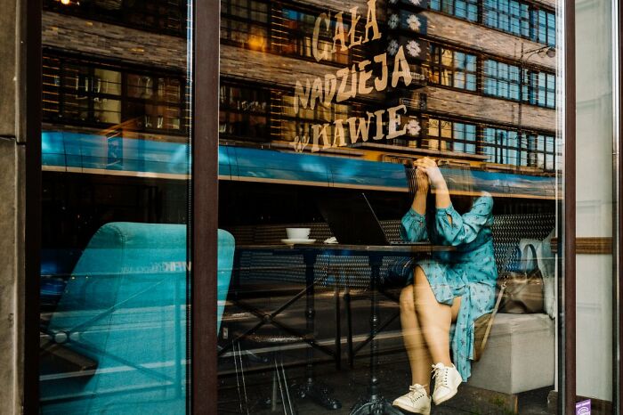 A woman sits inside a cafe, her reflection visible in the window, capturing a candid street photo for URBAN Photo Awards.