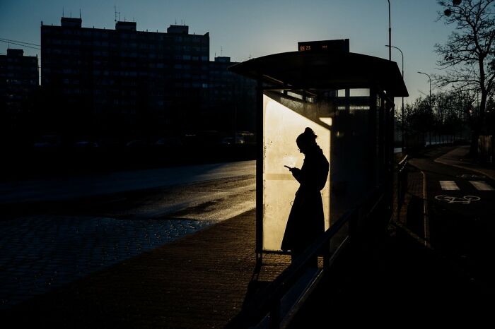 A silhouette of a person at a bus stop, bathed in strong backlight, reflecting a candid street photo for URBAN Photo Awards.
