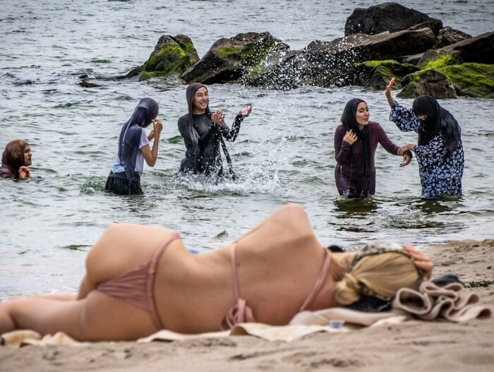 A candid street photo contrasting women swimming in modest attire with a woman sunbathing in a bikini, from URBAN Photo Awards.