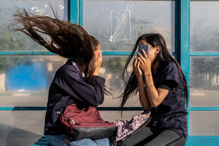 A candid street photo showing two young women at a bus stop with wind blowing their hair, featured in the URBAN Photo Awards.
