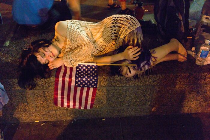 Candid street photo of a person lying on the ground next to a small American flag at an URBAN gathering.