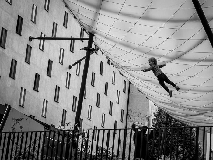 Candid street photo in black and white of a child on a large net above a man, an URBAN photo awards entry.