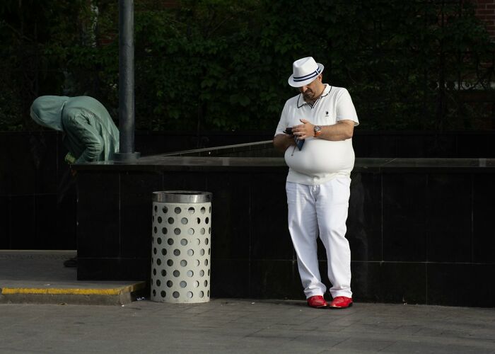 Candid street photo of a man in a white outfit and hat looking at his phone next to a bin, an URBAN photo awards entry.