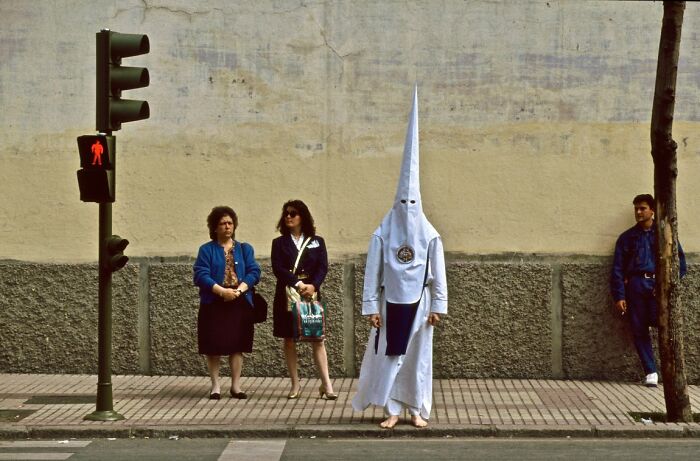 A person in a tall, pointed white hood stands on a sidewalk next to two women, a unique candid street photo from the URBAN Photo Awards.