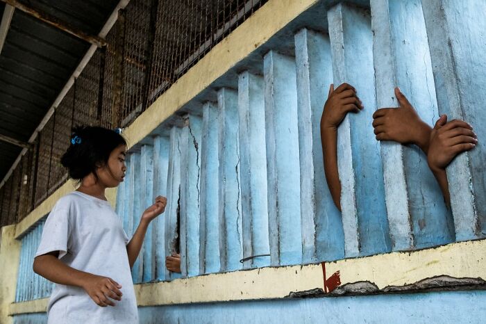A young girl looks toward hands reaching out from behind blue concrete bars, a poignant moment captured for the URBAN Photo Awards.