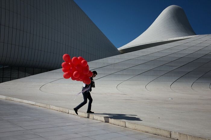 A man carries a large bunch of red balloons past a modern, curved building, a striking image from the URBAN Photo Awards.