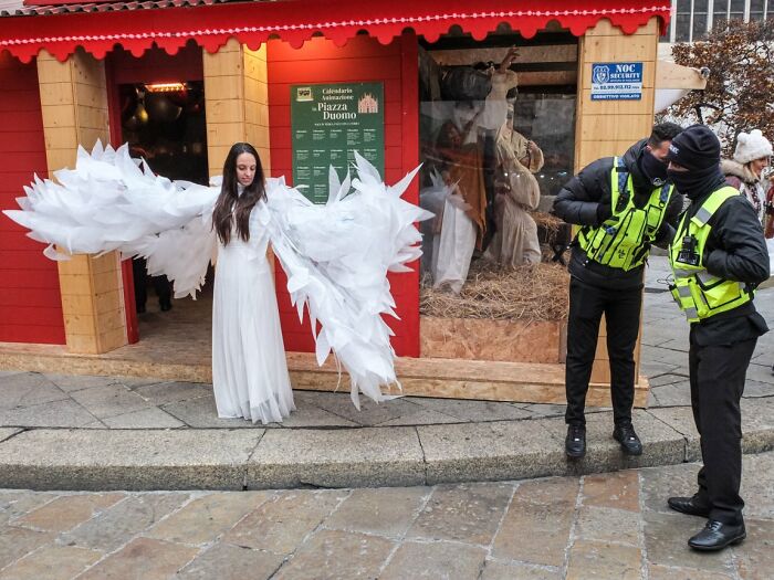 A woman in large white angel wings stands in front of a holiday display, part of the URBAN Photo Awards candid street photos.