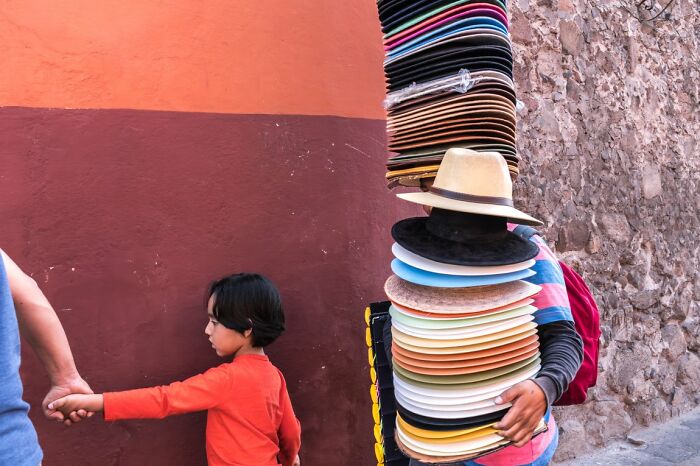 A street vendor carries a towering stack of hats while a child walks beside an adult, captured for URBAN Photo Awards.