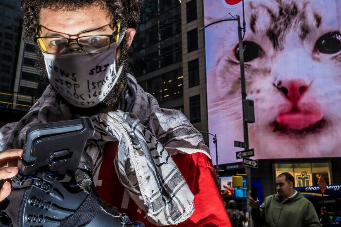 A man in a face mask holding a camera, with a giant cat image on a screen in the background, a Candid Street Photo.