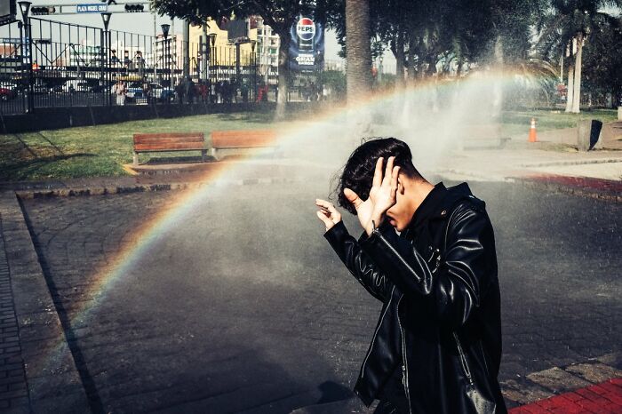A person in a black leather jacket covers their face as a rainbow forms from a street sprinkler in a candid street photo.