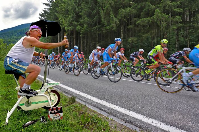 A man on a stationary bike holds out a bottle to passing cyclists during a race, a dynamic candid street photo.
