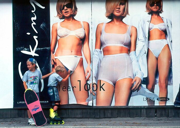 Two young boys, one with a skateboard and one on rollerblades, stand in front of a billboard with models in lingerie, a candid street photo.