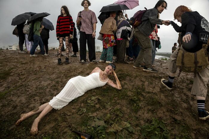 A person in a white dress lies in mud while others stand around under umbrellas on a rainy day, captured in a candid street photo.