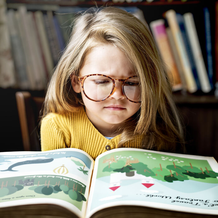 A young girl with glasses intently reading a picture book, highlighting childhood beliefs and imagination.