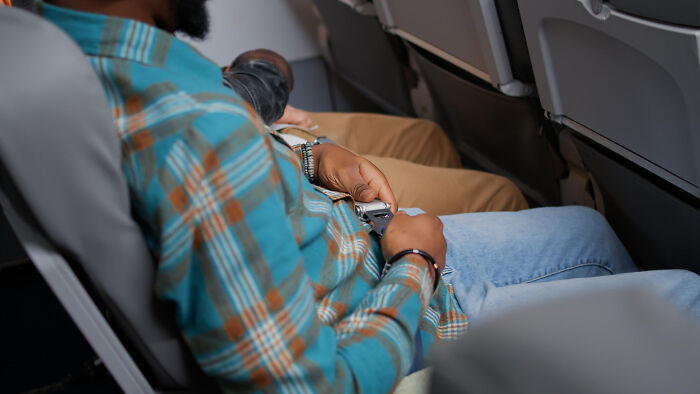 A man in a plaid shirt buckles his seatbelt on a plane, showing the space under a seat.