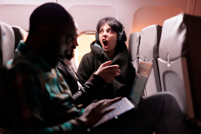 A woman wearing headphones yawns while an entitled man argues about seat space.