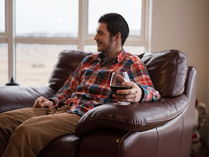 Man holding glass of red wine on leather couch reflecting on drinking problem Man holding glass of red wine on leather couch reflecting on drinking problem