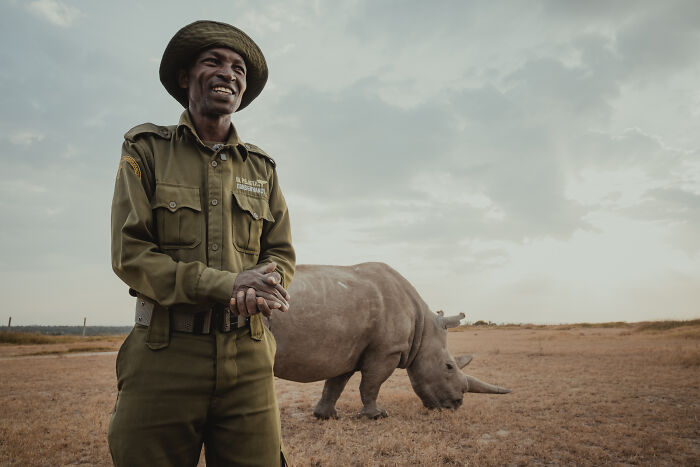 A smiling protector of the last northern white rhinos, in uniform, with a rhino grazing in the background.