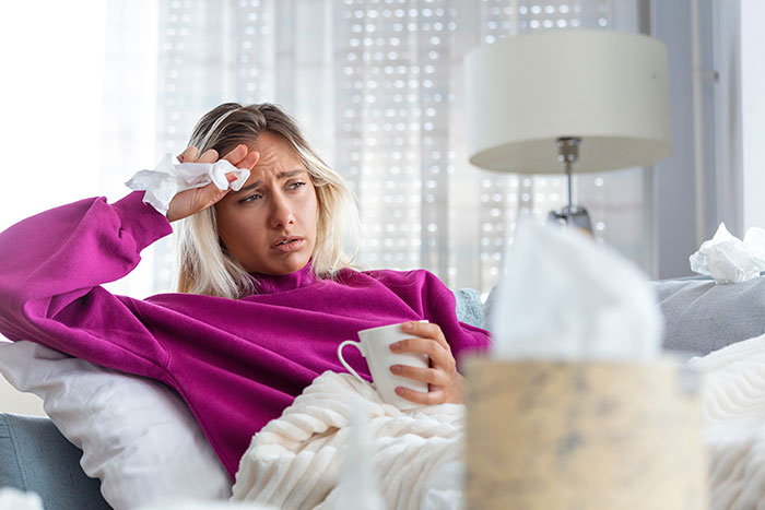 Woman feeling ill from allergy after raking, resting on a couch with tissues and a mug, refusing to cook for husband.