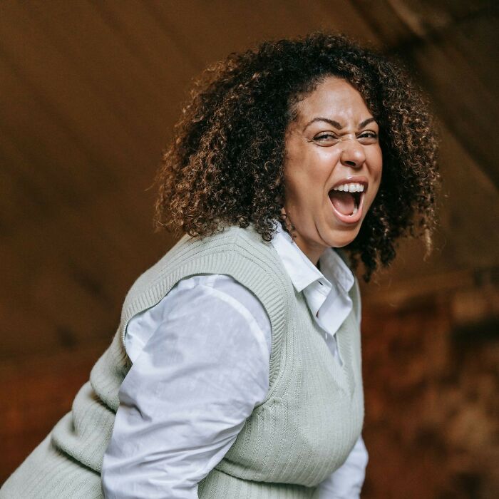 A woman with curly hair laughs loudly, expressing a funny everyday inconvenience.
