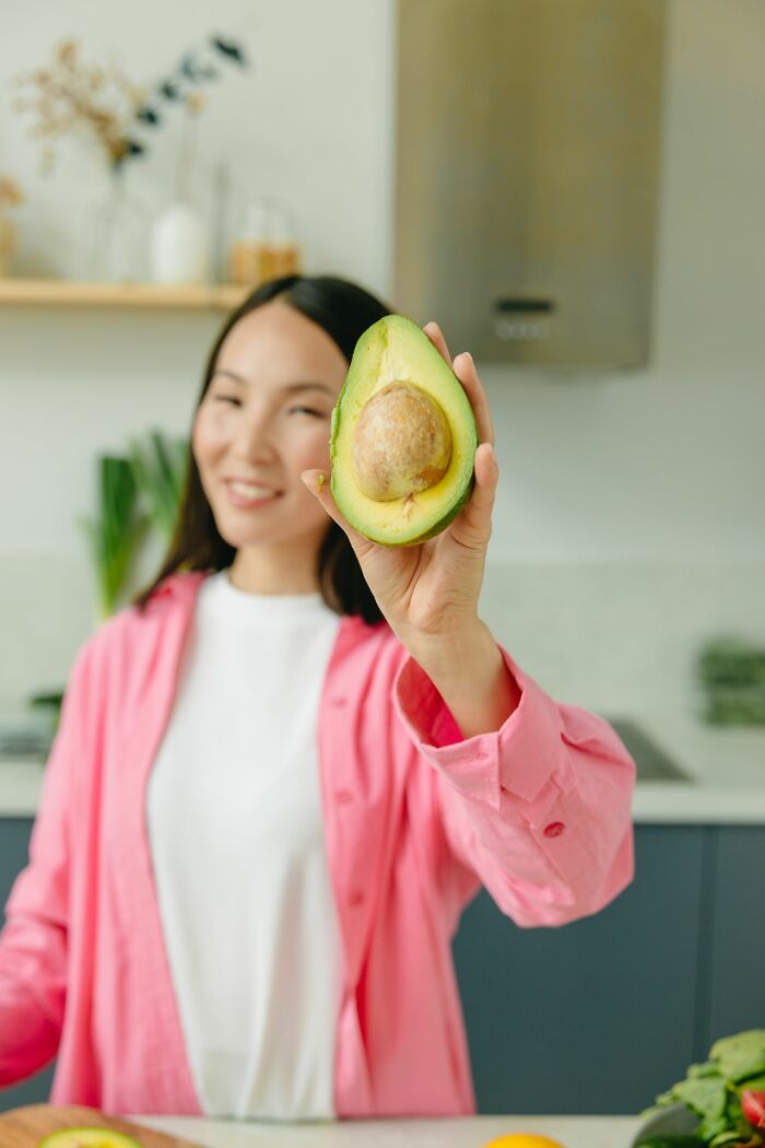 A smiling woman holding up half an avocado in a kitchen, a humorous take on everyday inconveniences.