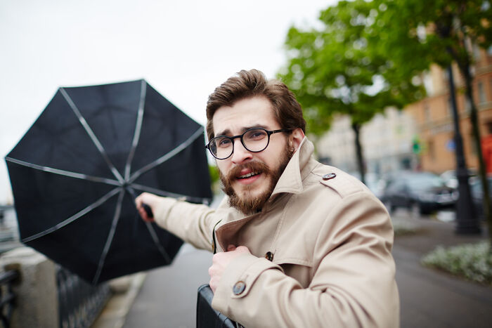 A man with a beard and glasses struggles with an inverted umbrella on a windy day, a common inconvenience.