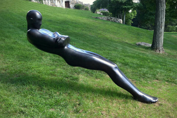 A baffling image of a black metallic human-like statue levitating diagonally over green grass on a sunny day.