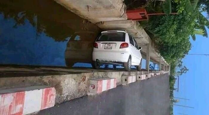 A white car precariously balanced on a narrow ledge next to a canal, a baffling image.