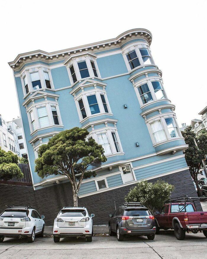 A baffling image of a blue building appearing to lean precariously on a hill, defying physical sense, with cars parked below.