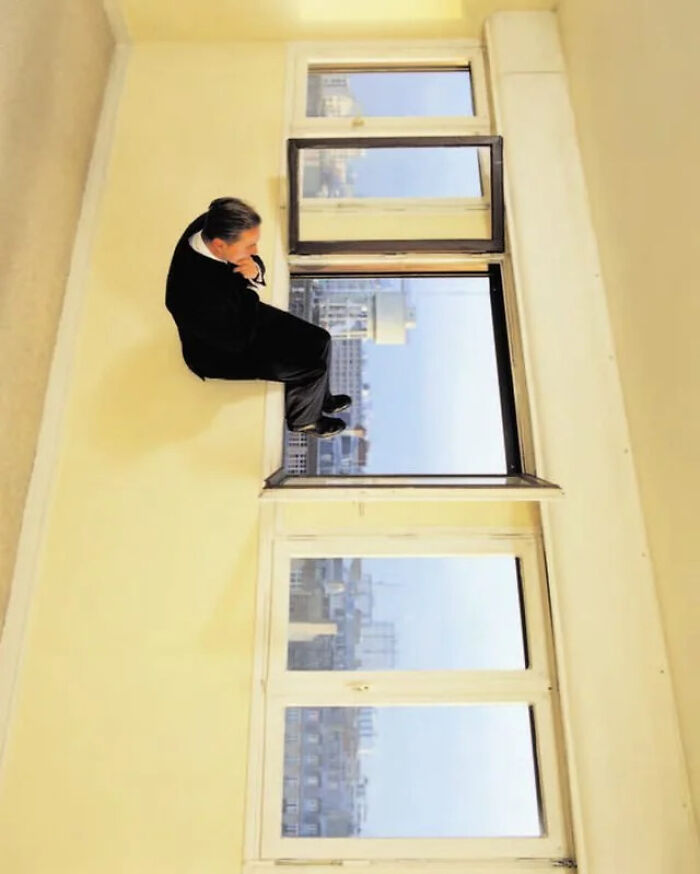A baffling image of a man appearing to sit on a window, defying physical sense, with a city view outside.