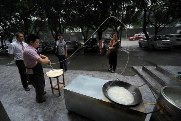 A baffling image of a man stretching dough into a long noodle over a large wok outdoors, a scene that makes no physical sense.