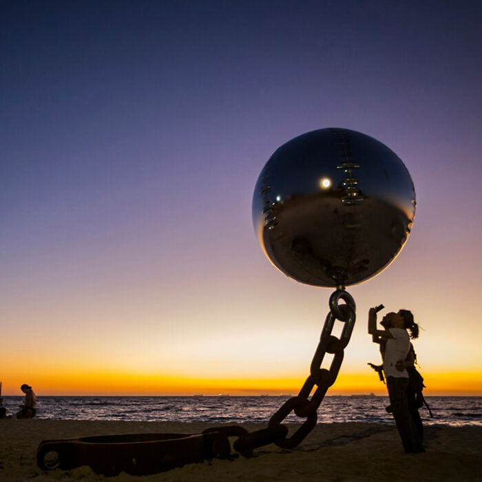 A giant chain and ball sculpture on a beach at sunset, forming a baffling image against the vibrant sky.