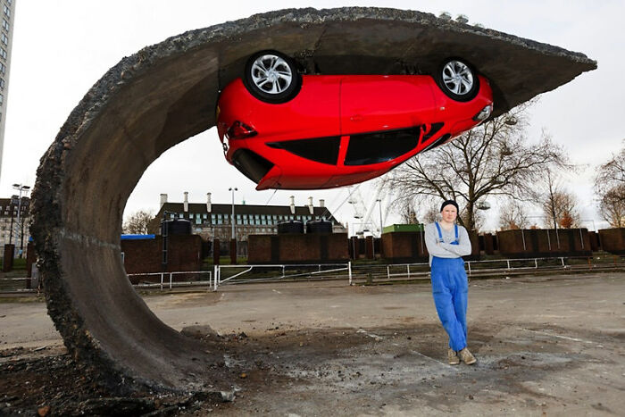 A baffling image of a man standing next to a red car suspended upside down under a curved concrete slab, defying physical sense.