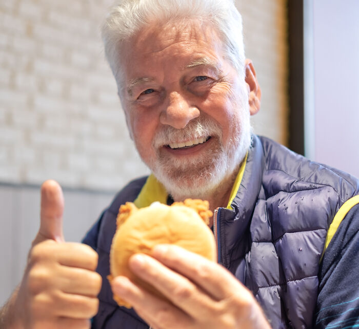 An elderly man smiling and giving a thumbs up while holding a burger, showing no pain reactions.