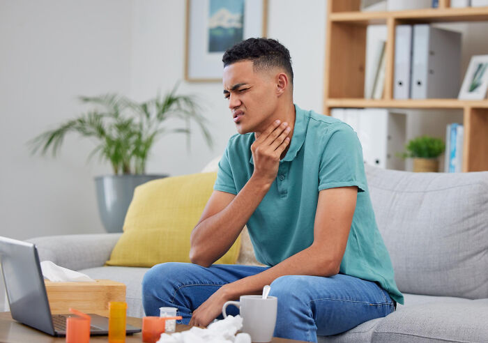 Young man on a couch touching his sore throat, surrounded by tissues and medicine, indicating serious issues.