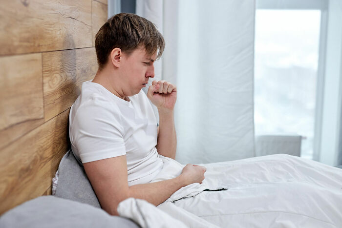 A man in bed coughs into his fist, representing a seemingly innocent symptom that could hide serious medical issues.