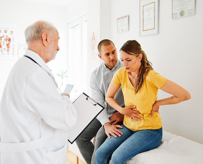 A medical professional consults with a couple; the woman clutches her stomach, showing signs of serious issues.