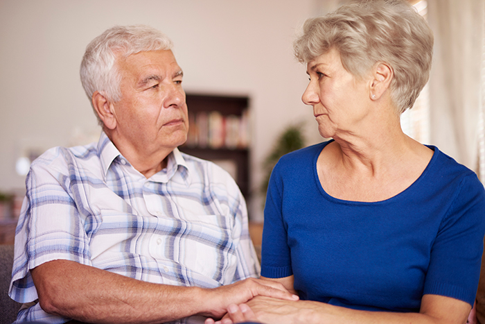 An older couple looking concerned while sitting together, representing tired hosts during family gatherings. An older couple looking concerned while sitting together, representing tired hosts during family gatherings.