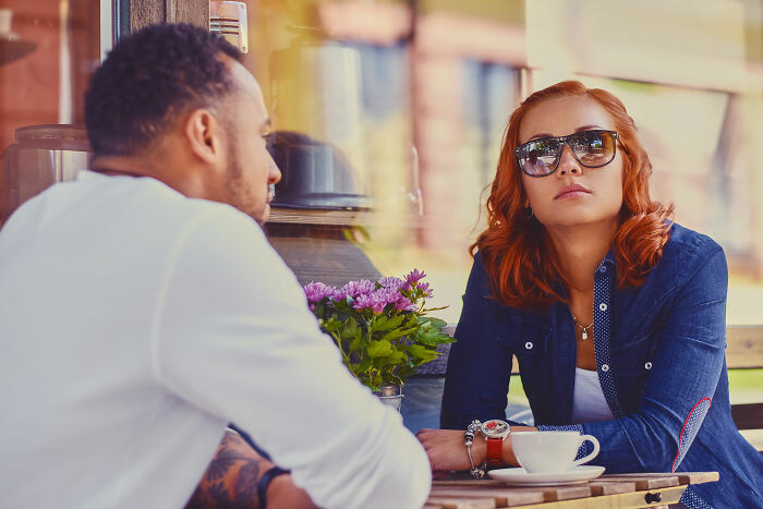 A man and a woman with red hair and sunglasses sitting at a cafe table, possibly discussing dating red flags.