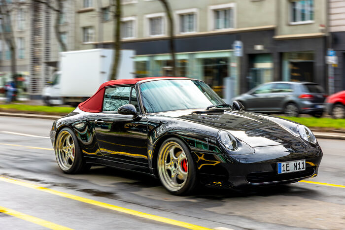 A black luxury sports car with a red convertible top drives on a city street, one of the surprising things wealthy people buy.