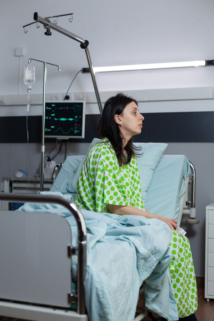 A woman in a hospital gown sitting on a bed, experiencing extreme illness and sickness.