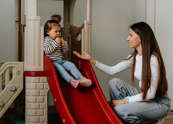A nanny playing with a young child on a red slide