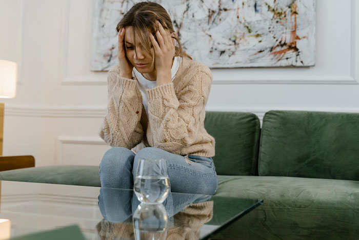 A stressed and anxious nanny sitting on a green couch holding her head after finding a hidden AirTag.