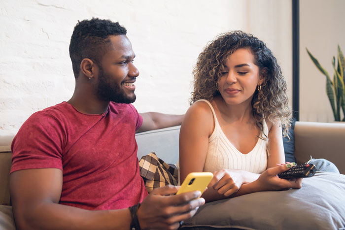 Couple sitting on couch, man holding phone and woman holding remote, discussing obsession topic