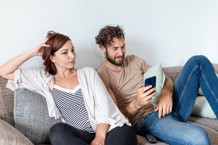 A young couple lounging on a couch, with the man looking at his phone, related to parents giving free housing.