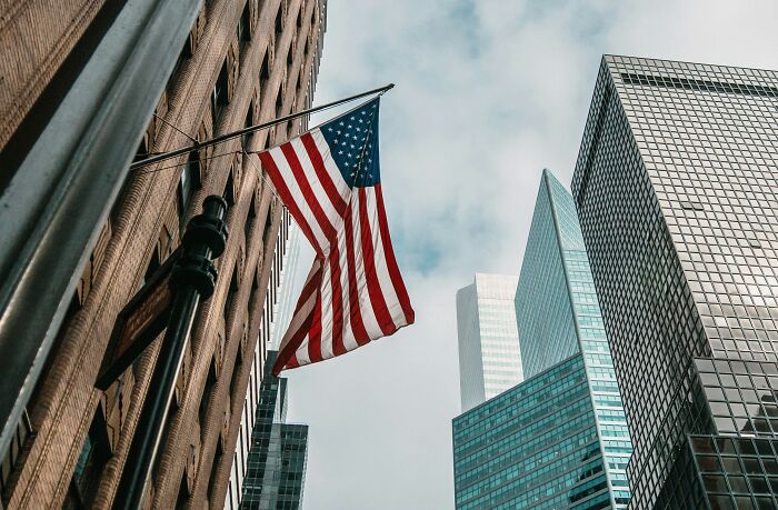 American flag waving between modern buildings, reflecting a peaceful sky.