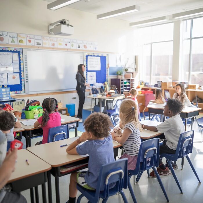 A teacher stands in front of a class of young children at their desks, embodying a childhood belief.