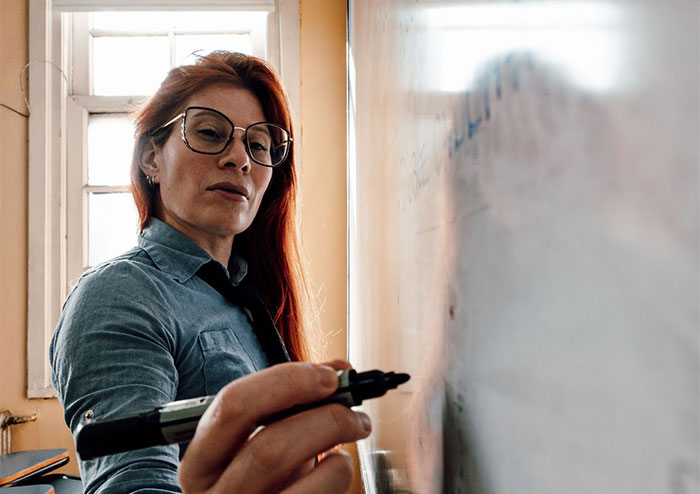A teacher with red hair and glasses looks intensely while writing on a whiteboard. A teacher with red hair and glasses looks intensely while writing on a whiteboard.