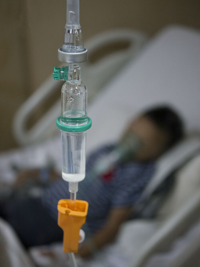 A close-up of an IV drip in a hospital, with a blurred patient on a bed with an oxygen mask in the background, showing medical pain reactions.